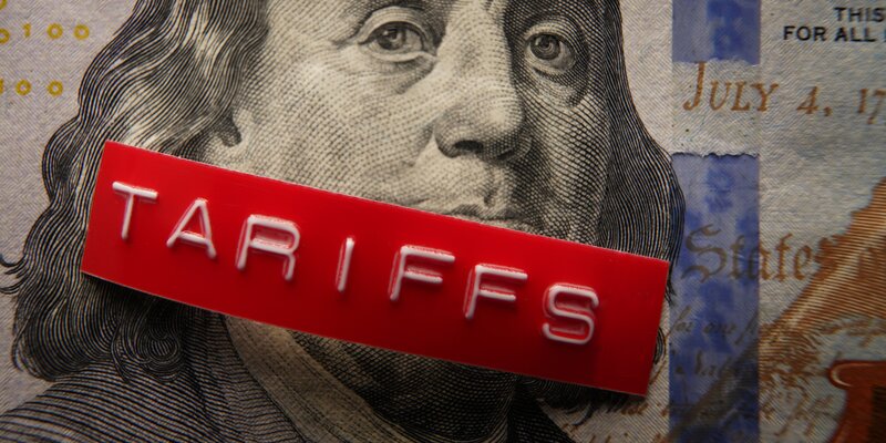 Close-up of a hundred-dollar bill with a red rectangular label featuring raised white letters spelling out tariffs and placed diagonally across the portrait.